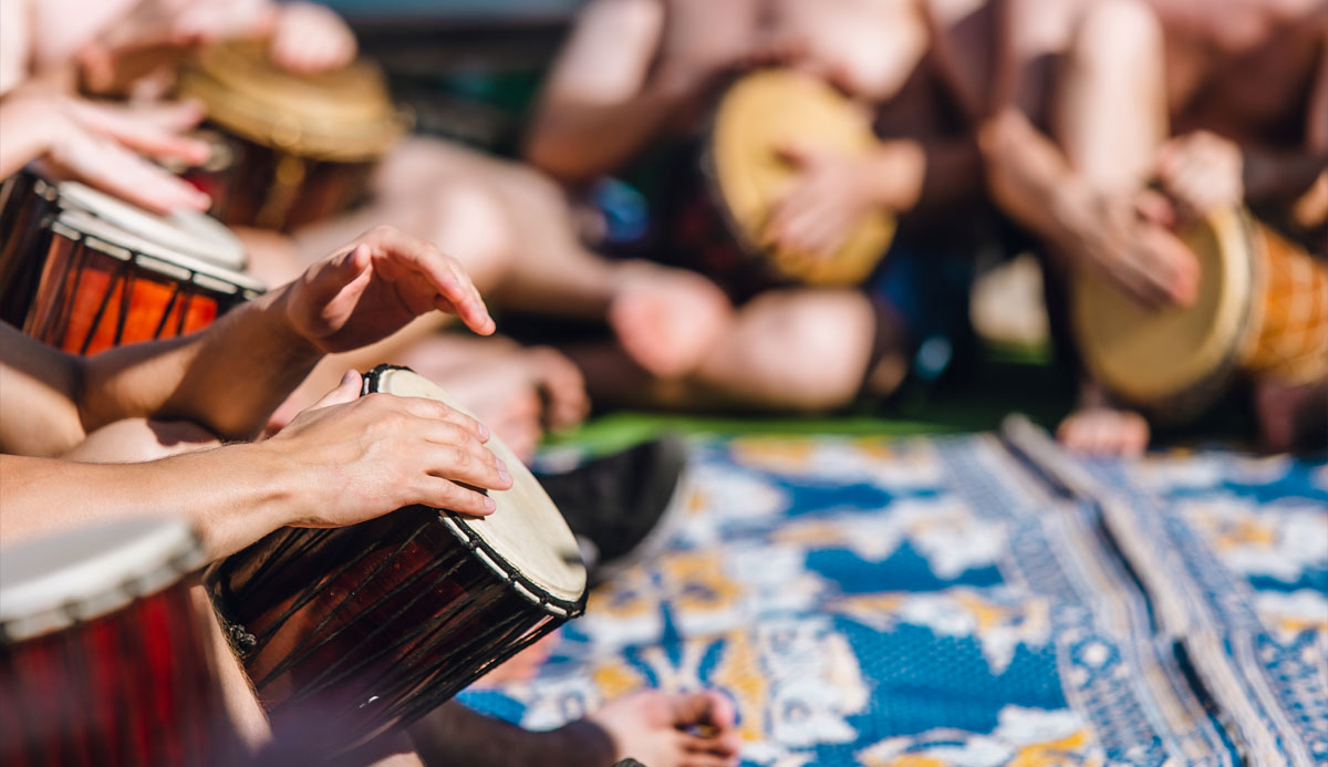 Close up of various people's hands playing drums in a drum circle sitting on a vibrant patterned blanket