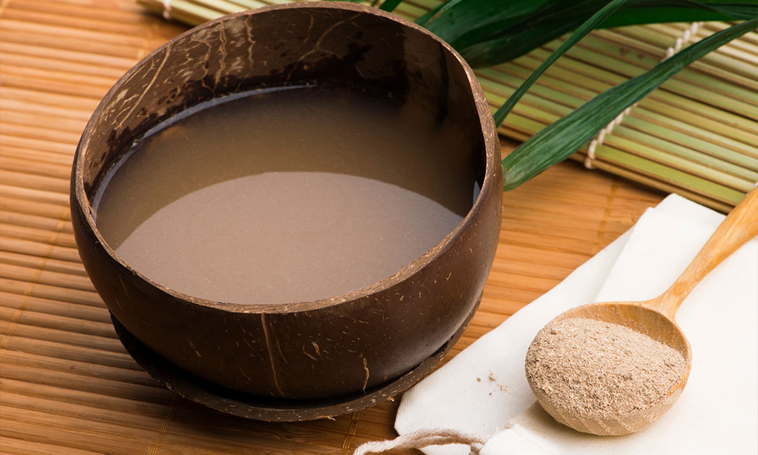 A wooden coconut bowl full of kava on a bamboo tray with a wooden spoon full of kratom powder