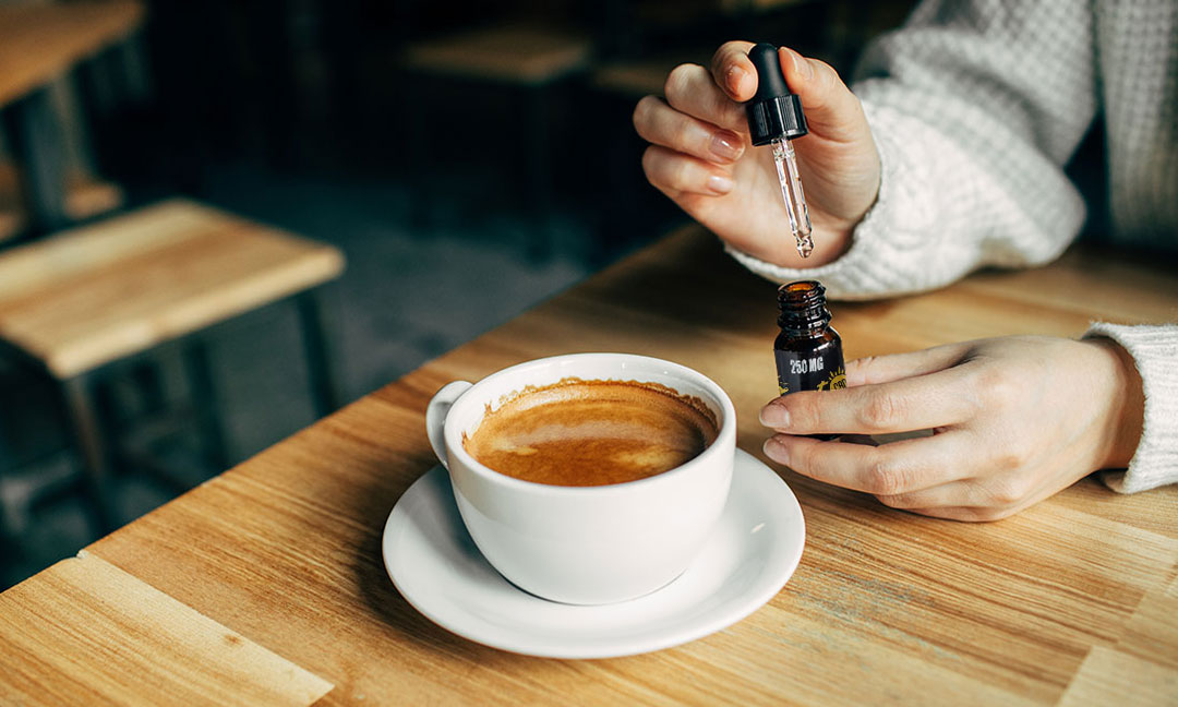 Close up of woman's hands holding a CBD oil dropper sitting in front of a mug of coffee